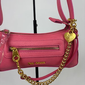 May include: Pink shoulder bag with a gold-tone zipper and hardware. The bag features a gold chain and a heart-shaped charm. The brand name "Juicy Couture" is visible on the front of the bag. The bag has a textured finish.