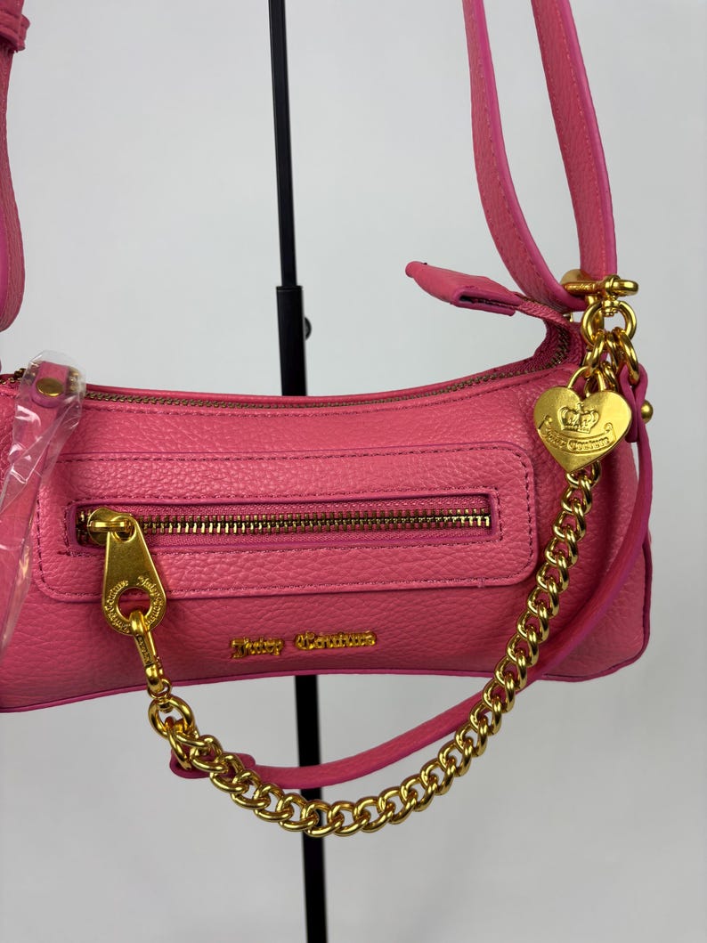 May include: Pink shoulder bag with a gold-tone zipper and hardware. The bag features a gold chain and a heart-shaped charm. The brand name "Juicy Couture" is visible on the front of the bag. The bag has a textured finish.