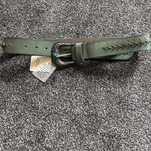 May include: A teal-colored belt with a decorative silver buckle and a turquoise accent. The belt features stitched detailing on the end and small silver studs. A tag is attached to the belt.