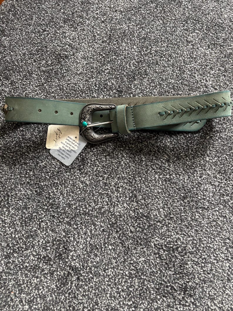 May include: A teal-colored belt with a decorative silver buckle and a turquoise accent. The belt features stitched detailing on the end and small silver studs. A tag is attached to the belt.