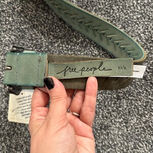 May include: A distressed teal leather belt with a stitched design and a metal buckle. The belt features the brand name "free people" in cursive script. The size is XS/S.
