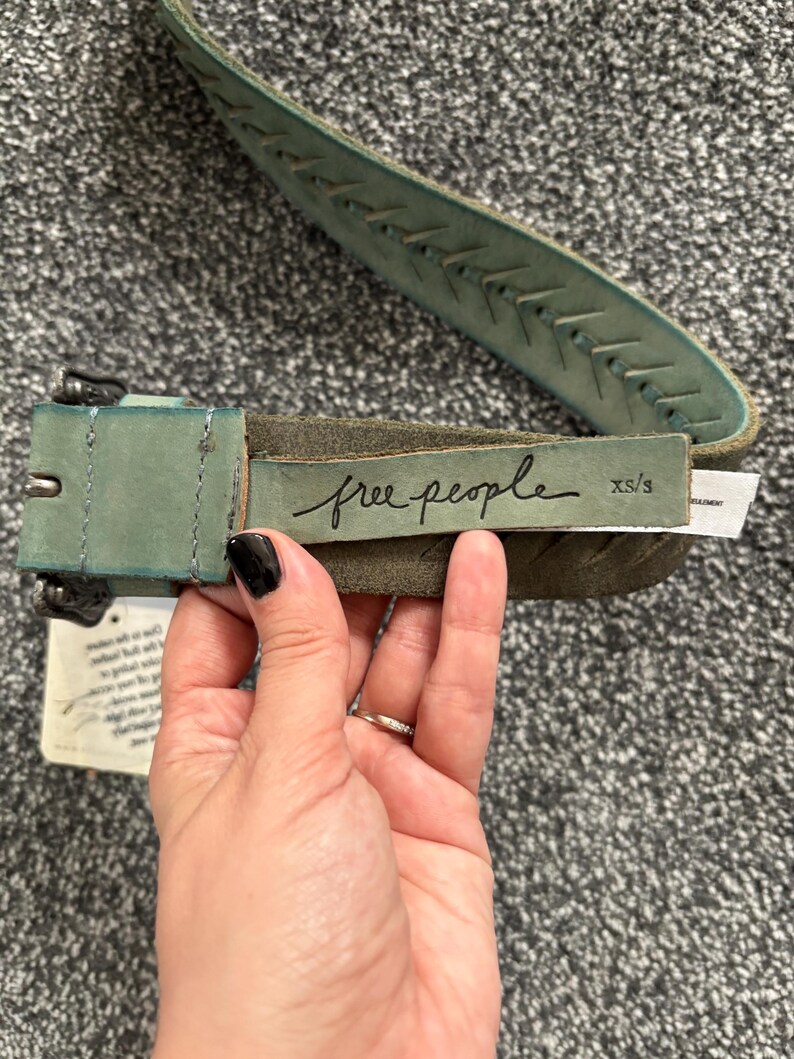 May include: A distressed teal leather belt with a stitched design and a metal buckle. The belt features the brand name "free people" in cursive script. The size is XS/S.