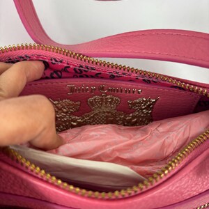 May include: Pink leather crossbody bag with a gold-tone chain strap and zipper. The interior features a leopard print lining and a silver Juicy Couture logo. The bag is open, revealing tissue paper.