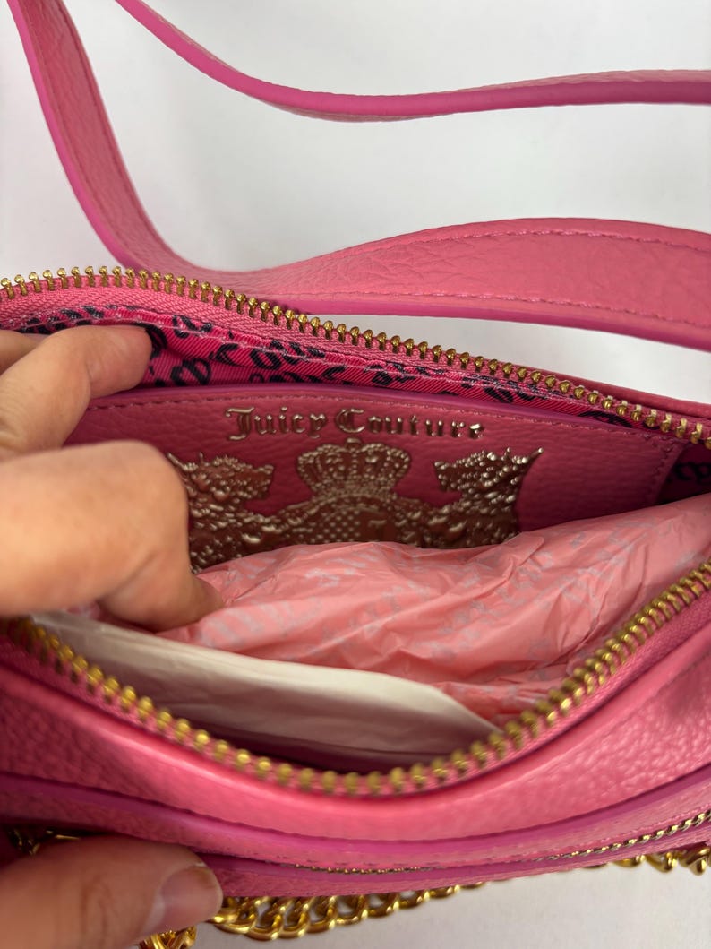 May include: Pink leather crossbody bag with a gold-tone chain strap and zipper. The interior features a leopard print lining and a silver Juicy Couture logo. The bag is open, revealing tissue paper.