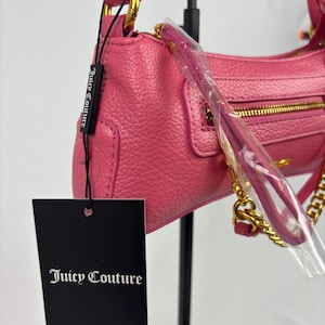 May include: A pink leather shoulder bag with gold-tone hardware, including a chain strap. The bag features a zippered closure and a small pocket on the front. A black tag with the "Juicy Couture" logo is attached.