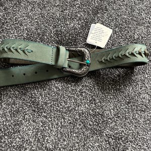 May include: A sage green leather belt with a decorative silver buckle and turquoise accent. The belt features stitched detailing along the edges and a classic buckle closure. A tag is attached to the belt.