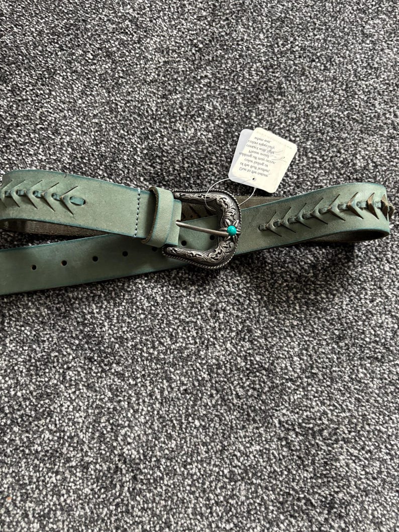 May include: A sage green leather belt with a decorative silver buckle and turquoise accent. The belt features stitched detailing along the edges and a classic buckle closure. A tag is attached to the belt.
