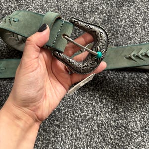 May include: A teal leather belt with a decorative silver buckle. The buckle features intricate floral designs and a small turquoise stone. The belt has a stitched design along the edges.