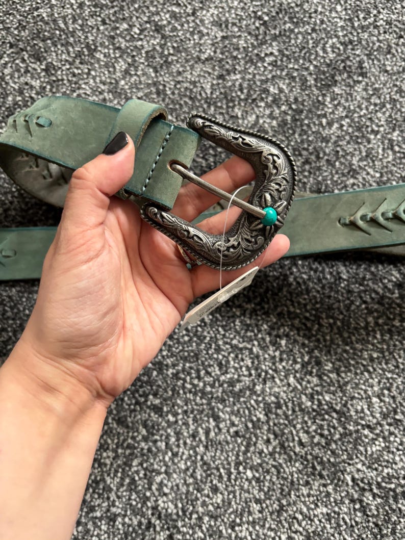 May include: A teal leather belt with a decorative silver buckle. The buckle features intricate floral designs and a small turquoise stone. The belt has a stitched design along the edges.