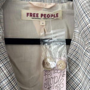 May include: A blazer with a plaid pattern in shades of gray, white, and brown. The interior lining is a light beige color. The label reads "FREE PEOPLE". Two buttons are attached to a pink tag that reads "Free People".