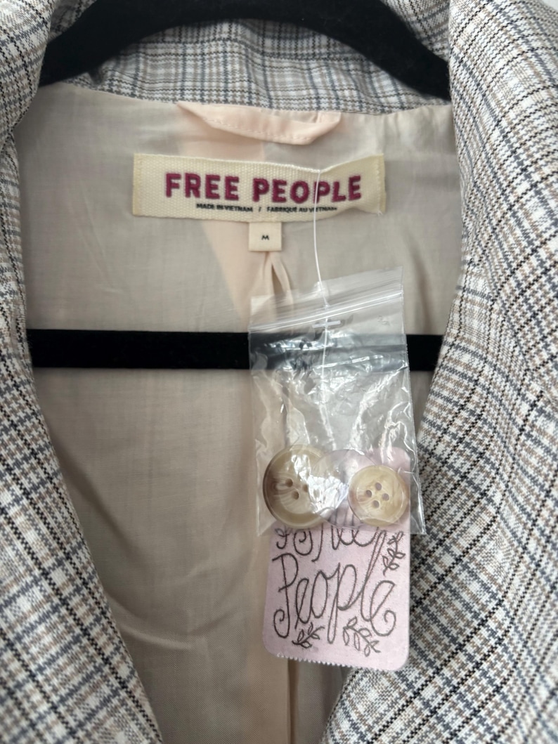 May include: A blazer with a plaid pattern in shades of gray, white, and brown. The interior lining is a light beige color. The label reads "FREE PEOPLE". Two buttons are attached to a pink tag that reads "Free People".