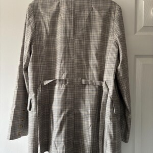 May include: A tan and white plaid blazer with a notched collar, long sleeves, and a tie belt. The blazer has a button closure on the sleeves and a tailored fit. The back of the blazer is visible, hanging on a white door.