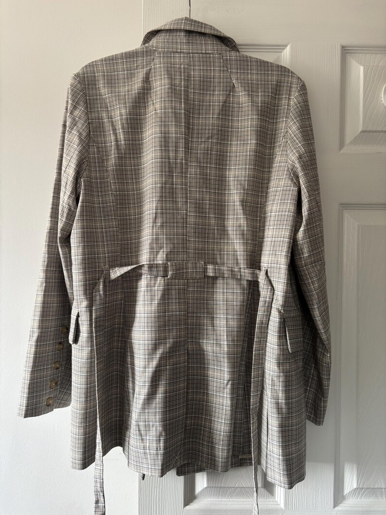 May include: A tan and white plaid blazer with a notched collar, long sleeves, and a tie belt. The blazer has a button closure on the sleeves and a tailored fit. The back of the blazer is visible, hanging on a white door.