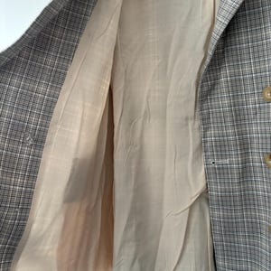 May include: A plaid blazer in shades of gray, black, and beige, with a light beige lining. The blazer has a classic check pattern and a button closure. The interior lining is visible, showing the tailored construction of the garment.