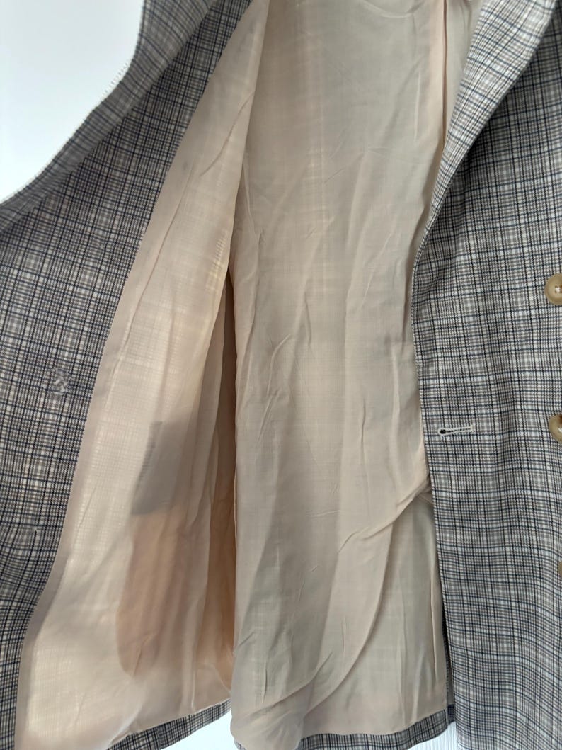 May include: A plaid blazer in shades of gray, black, and beige, with a light beige lining. The blazer has a classic check pattern and a button closure. The interior lining is visible, showing the tailored construction of the garment.
