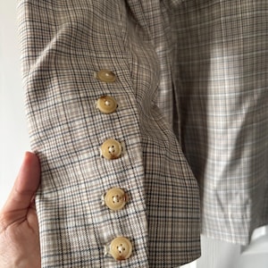 May include: A tan and brown plaid blazer with five round, light-colored buttons on the sleeve. The blazer has a classic plaid pattern and a tailored fit, suitable for various occasions.