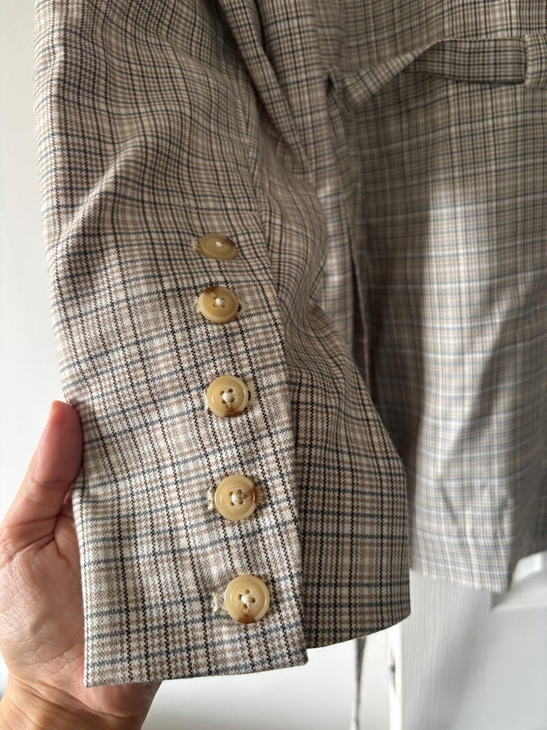 May include: A tan and brown plaid blazer with five round, light-colored buttons on the sleeve. The blazer has a classic plaid pattern and a tailored fit, suitable for various occasions.