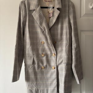 May include: A double-breasted blazer in a plaid pattern, featuring a neutral color palette of beige, gray, and white. The jacket has four large, round, light-colored buttons and two front pockets. The label reads "FREE PEOPLE".