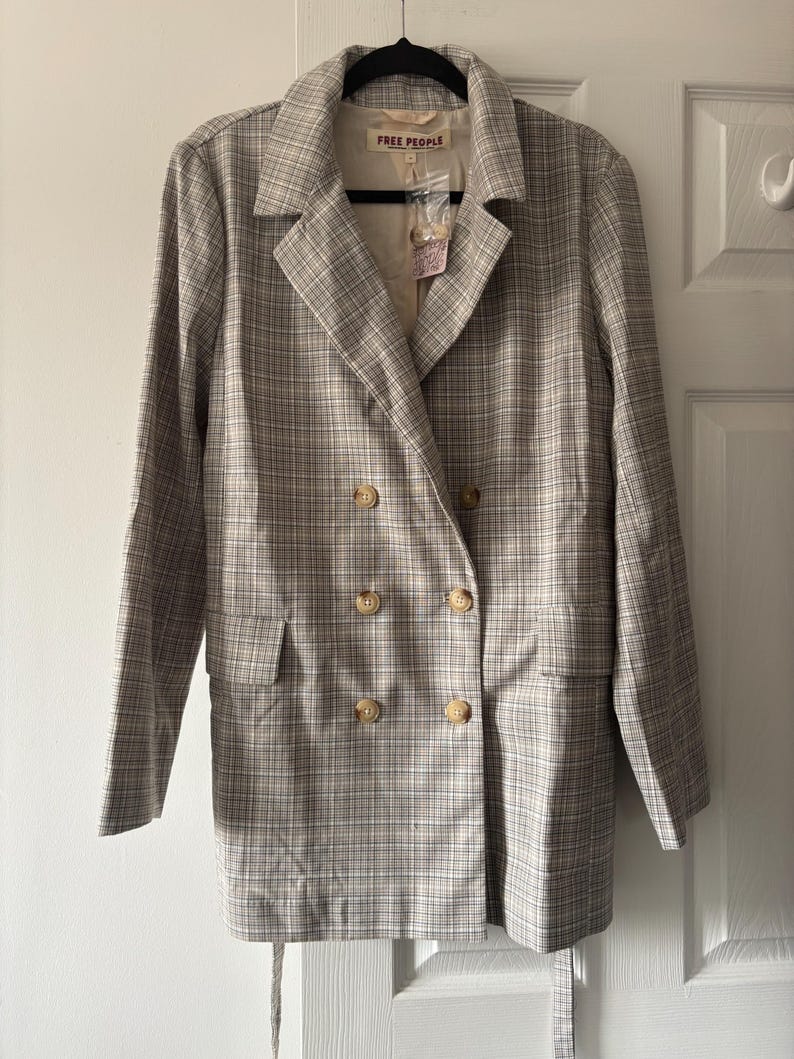 May include: A double-breasted blazer in a plaid pattern, featuring a neutral color palette of beige, gray, and white. The jacket has four large, round, light-colored buttons and two front pockets. The label reads "FREE PEOPLE".