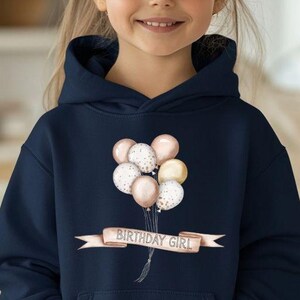 May include: Navy blue hooded sweatshirt featuring a balloon graphic and the text "BIRTHDAY GIRL". The graphic includes a cluster of balloons in shades of pink, white, and gold. The sweatshirt has a front pocket and a drawstring hood.