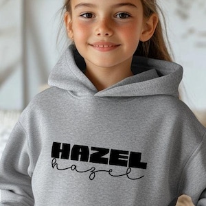 May include: A gray hooded sweatshirt with the name "HAZEL" printed in black block letters above a cursive "hazel". The sweatshirt has a front pocket and is being worn by a person wearing blue jeans.