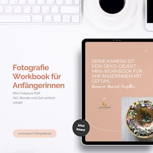 Op de afbeelding: Een digitale tablet toont een fotografie werkboek getiteld "Fotografie Workbook für Anfängerinnen". De tekst is in het Duits. De tablet ligt op een wit oppervlak met een toetsenbord en een koffiekopje.