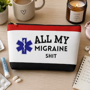 May include: A white, red, and black zippered cosmetic bag with the text "ALL MY MIGRAINE SHIT" and a blue medical symbol. The bag is surrounded by medication, a candle, and a notebook labeled "Migraine Kit".