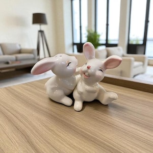 May include: Two white ceramic bunny figurines with pink inner ears and smiling faces. One bunny is sitting, and the other is leaning in, appearing to touch the other. The figurines are on a wooden surface.