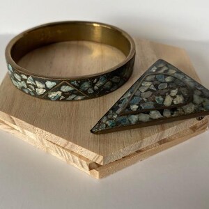 Può includere: Un braccialetto a cerchio in ottone e una spilla triangolare con intarsi a mosaico di pietre blu, verdi e bianche. I gioielli poggiano su un piccolo blocco di legno chiaro.