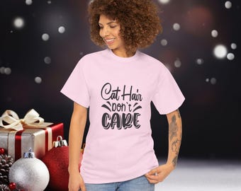 Pode incluir: Camiseta rosa claro com o texto preto "Cat Hair Don't Care". A pessoa está usando a camiseta. No fundo, decorações de Natal, como bolas e presentes embrulhados.
