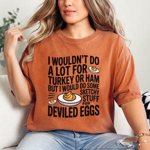 May include: Burnt orange t-shirt with the text "I wouldn't do a lot for turkey or ham but I would do some sketchy stuff for deviled eggs." The shirt features a graphic of deviled eggs. The word "Yam" is in the bottom right corner.