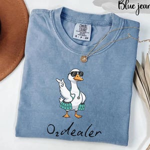May include: Blue jean colored t-shirt with a cartoon goose wearing sunglasses and a stethoscope. The goose is giving a hand gesture. The text "O2dealer" is written in cursive at the bottom of the shirt.
