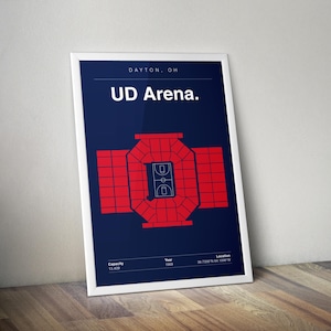 Op de afbeelding: Een ingelijste poster met een rood-blauwe afbeelding van de UD Arena in Dayton, OH. De poster bevat de tekst "UD Arena" en details over de capaciteit, het jaar en de locatie van de arena. De poster leunt tegen een muur.