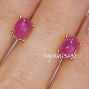 May include: A pair of gold stud earrings, each with an oval ruby gemstone. The rubies are a deep pinkish-red colour, set in a gold bezel with small gold accents. The earrings are displayed on a hand with silver glitter nail polish. The text "myasajewels" is visible.