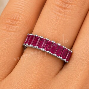 Peut inclure: Une bague argentée ornée d'une rangée de pierres précieuses rectangulaires taille baguette d'un rouge rose profond, fixées dans un sertissage à griffes partagées. L'anneau est fin et métallique, présentant une série de pierres vibrantes.