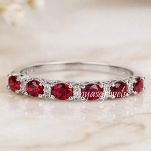 May include: A silver ring featuring a series of oval-cut, vibrant red gemstones, interspersed with smaller, clear stones. The ring is set against a soft, neutral background, highlighting the sparkle of the gemstones. The text "myascjewels" is visible on the ring.