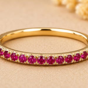 May include: A gold band ring with a continuous row of vibrant red gemstones. The ring's polished gold setting provides a striking contrast to the bright red stones. The ring is set against a neutral background.