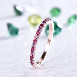 May include: Rose gold ring with a row of alternating red and purple gemstones. The ring is set against a blurred background with green and yellow accents, creating a colorful and elegant display.