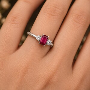 May include: A silver-toned ring featuring a central rectangular emerald-cut deep red gemstone flanked by two smaller round brilliant-cut clear stones. The band is smooth and polished, holding the three stones in a prong setting arrangement.