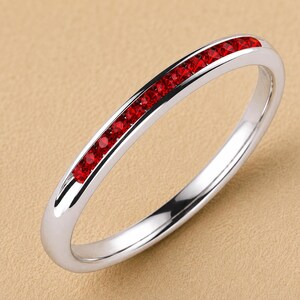 Puede incluir: Un anillo de banda en tono plateado pulido que presenta una fila de pequeñas piedras circulares de color rojo vibrante engastadas en canal a lo largo de la mitad superior de la superficie exterior.