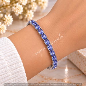 Può includere: Un bracciale tennis caratterizzato da una serie di pietre preziose azzurre dal taglio ovale, incastonate in una fascia di metallo color argento lucido. Il bracciale è indossato al polso, mostrando la disposizione uniforme delle pietre.