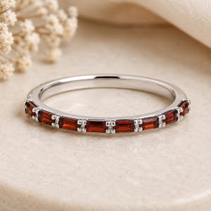 May include: A silver-toned band ring featuring a series of rectangular, deep red garnet gemstones set in a channel style. The stones are arranged horizontally around the front half of the band, separated by small, polished silver-toned metal spacers.