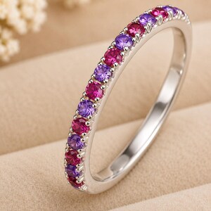 May include: A silver-toned band ring featuring a row of alternating round-cut fuchsia pink and violet purple gemstones set along the top half of the metal band. The ring has a polished finish and a slim profile with a continuous channel setting.