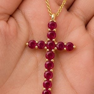 Puede incluir: Un colgante de cruz en oro con once rubíes redondos de color rojo intenso engastados en una montura de metal con garras. La cruz cuelga de una delicada cadena de oro tipo cuerda, mostrando un diseño simétrico con una piedra central.