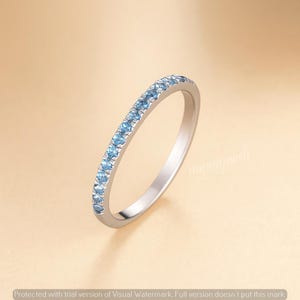 Könnte beinhalten: Ein silberner Ring mit einer Reihe kleiner, hellblauer Edelsteine. Der Ring ist vor einem weichen, neutralen Hintergrund platziert, der das Funkeln der Steine hervorhebt. Ein zartes Schmuckstück.