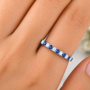 Puede incluir: Un anillo de banda en tono plateado que presenta un patrón alterno de zafiros azules de corte redondo y ópalos blancos de corte redondo engastados en una sola fila sobre la parte superior, creando un efecto brillante continuo.