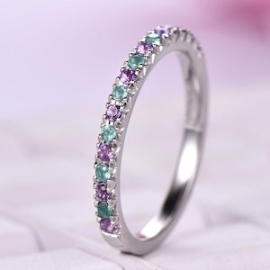 Może przedstawiać: Srebrny pierścionek z rzędem małych, kolorowych kamieni szlachetnych. Kamienie szlachetne naprzemiennie fioletowe i turkusowe, tworząc żywy i przyciągający wzrok design. Pierścionek na miękkim, rozmytym tle.