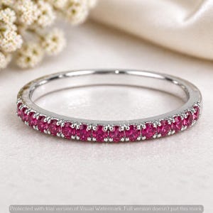 Könnte beinhalten: Ein silberner Bandring mit einer durchgehenden Reihe kleiner, leuchtend rosa Edelsteine. Das Metall des Rings hat eine polierte Oberfläche, und die Edelsteine sind eng aneinander gesetzt, was einen funkelnden Effekt erzeugt. Der Ring ist ein zartes Schmuckstück.