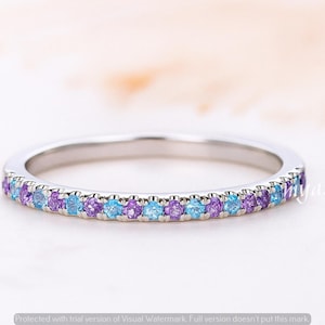 May include: A silver ring with a band of alternating purple and light blue gemstones. The ring is set against a soft, blurred background. The ring is a piece of jewelry.