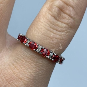 May include: A silver ring with a row of red gemstones and interspersed white gemstones. The ring is on a finger, with the brand name "myjsajewels" visible. The ring is set against a light blue background.